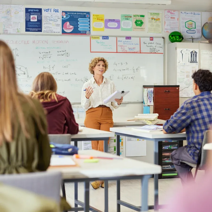 A teacher leads discussion at the front of a poster-lined classroom.