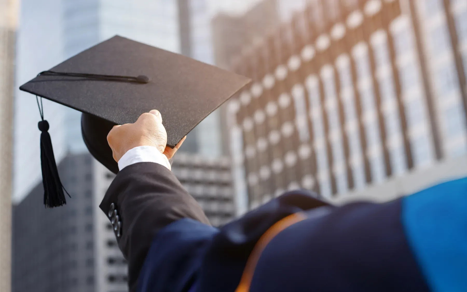 A graduate holds a mortarboard up against a city skyline.